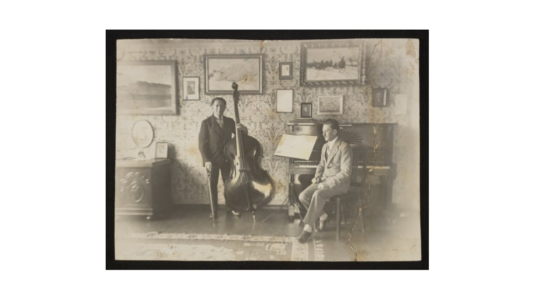 Czarno-biała fotografia przedstawia dwóch mężczyzn, jeden stoi z kontrabasem, a drugi siedzi obok pianina na stołku fortepianowym