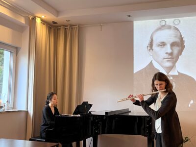 Fotografia przedstawia grającą flecistkę i pianistkę przy fortepianie na tle ekranu z wyświetlonym portretem mężczyzny (kompozytora)