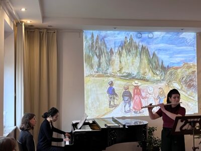 Zdjęcie przedstawia flecistkę i pianistkę podczas występu w Studiu Gallois, na tle ekranu, na którym wyświetlany jest dziecięcy rysunek. Obok pianistki widać osobę, która przewraca nuty podczas koncertu