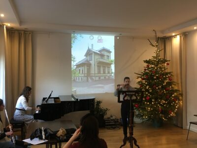 Studio Gallois - trwa koncert z udziałem flecistki i pianistki, które występują na tle ściany, na której wyświetlana jest fotografia budynku; obok flecistki stoi choinka z ozdobami świątecznymi.