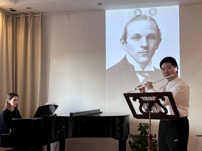 Zdjęcie z koncertu w ramach Kultura 3.0 przedsawia flecistę i pianistkę w Studiu Gallois, a za nimi ekran z wyświetlonym zdjęciem kompozytora