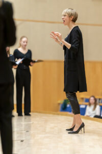 Na zdjęciu elegancko ubrana kobieta dyryguje chórem na estradzie - Marianna Majchrzak