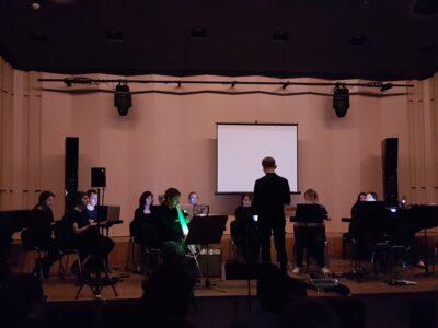 Zdjęcie przedstawia ciemną salę koncertową i czlonków LambdaEnsemble podczas koncertu w ramach Mulitmedialnej Majówki